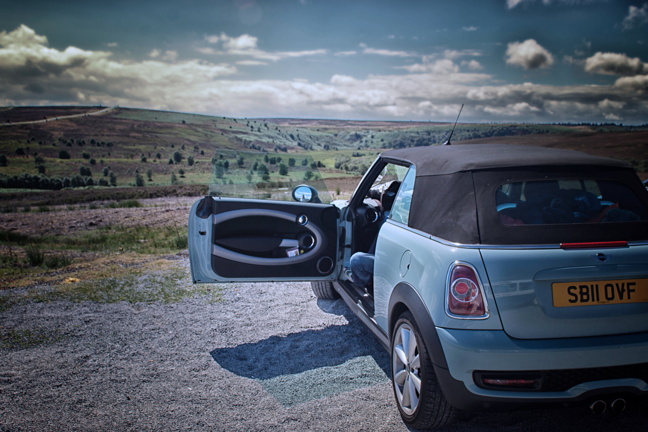 A BMW Mini Cooper convertible in a lay. The door is open and there are cumulus clouds on a bright summer's day.